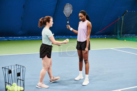 Téléchargez les photos : Portrait complet d'une entraîneuse de tennis travaillant avec une jeune femme noire pendant l'entraînement à la cour intérieure - en image libre de droit