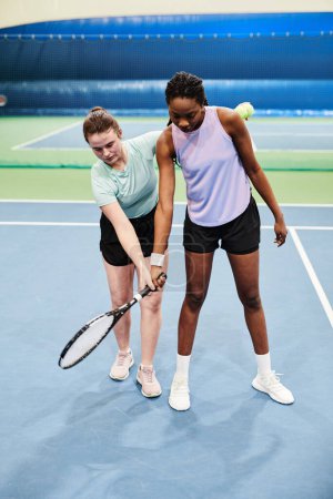 Téléchargez les photos : Portrait complet d'une entraîneuse travaillant avec une jeune femme noire pendant un entraînement de tennis au court intérieur - en image libre de droit