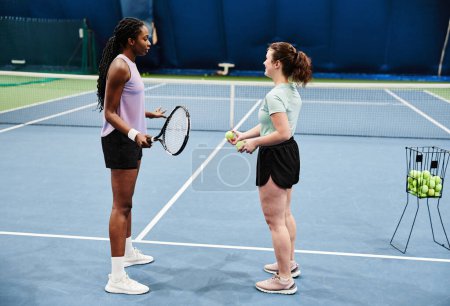 Téléchargez les photos : Portrait latéral d'une entraîneuse travaillant avec une jeune femme noire pendant un entraînement de tennis au court intérieur - en image libre de droit