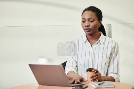 Téléchargez les photos : Portrait minimal de jeune femme noire utilisant un ordinateur portable tout en travaillant dans un immeuble de bureaux, espace de copie - en image libre de droit