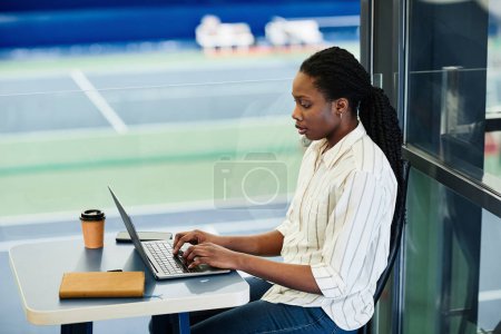 Foto de Retrato de vista lateral de una joven mujer negra que usa un portátil mientras trabaja en un centro de entrenamiento deportivo - Imagen libre de derechos