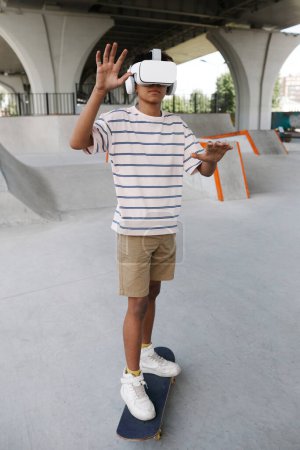 Foto de Retrato vertical de cuerpo entero de adolescente negro montando skate en VR auriculares al aire libre - Imagen libre de derechos