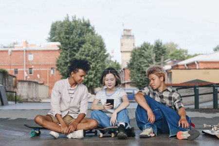 Téléchargez les photos : Portrait complet d'un groupe diversifié de trois locataires dans un skatepark à l'extérieur assis sur le sol et utilisant un environnement urbain smartphone - en image libre de droit