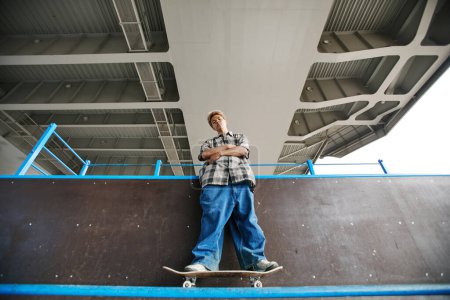 Téléchargez les photos : Portrait à angle bas d'un adolescent portant un pantalon baggy debout sur une planche à roulettes dans une zone urbaine et regardant une caméra, un espace de copie - en image libre de droit