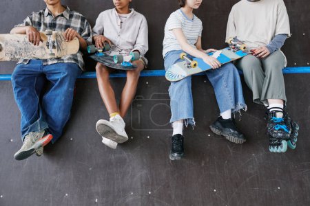 Foto de Diverso grupo de adolescentes sentados en la rampa en el parque de skate sección baja de los pies colgando, espacio de copia - Imagen libre de derechos