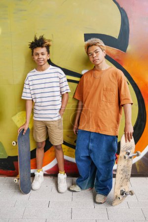 Téléchargez les photos : Portrait vertical de deux adolescents avec des planches à roulettes debout contre un mur de graffiti en vert et regardant la caméra - en image libre de droit