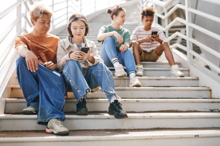 Photo for Airy full length shot of diverse teenagers using smartphones outdoors while sitting on metal stairs in sunlight - Royalty Free Image