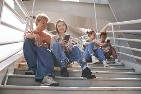 Photo for Sunlit full length shot of diverse teenagers using smartphones outdoors while sitting on metal stairs - Royalty Free Image