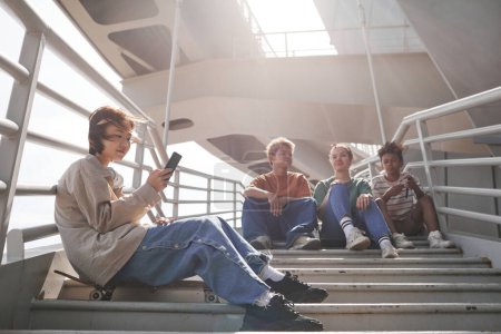 Téléchargez les photos : Plan aérien d'un groupe diversifié d'adolescents assis sur des escaliers en métal à l'extérieur en milieu urbain - en image libre de droit