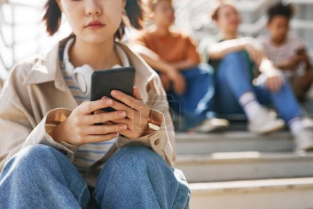 Foto de Primer plano de la adolescente sosteniendo el teléfono inteligente al aire libre mientras está sentado en escaleras de metal con un grupo de amigos en segundo plano, espacio para copiar - Imagen libre de derechos