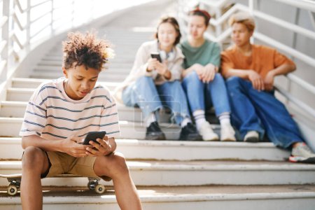 Foto de Retrato de un adolescente negro usando un teléfono inteligente al aire libre mientras está sentado en escaleras de metal con un grupo de amigos en segundo plano, espacio para copiar - Imagen libre de derechos