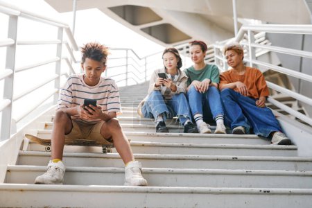 Foto de Retrato de larga duración de adolescente usando teléfono inteligente al aire libre mientras está sentado en escaleras de metal con un grupo de amigos en el fondo, espacio de copia - Imagen libre de derechos