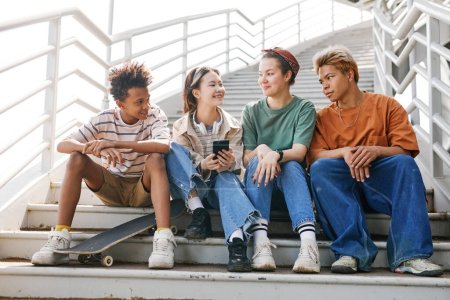 Foto de Retrato de larga duración de diverso grupo de adolescentes sentados en escaleras de metal al aire libre en el entorno urbano todo sonriendo y charlando - Imagen libre de derechos