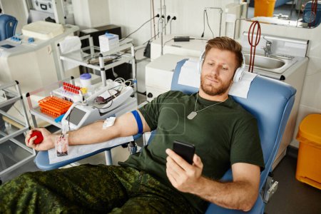 Téléchargez les photos : Portrait en grand angle d'un militaire donnant du sang alors qu'il était assis dans une chaise au centre de don de sang et regardant des vidéos sur smartphone, copiez l'espace - en image libre de droit