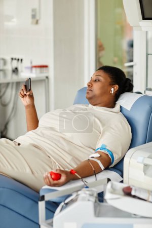 Foto de Retrato vertical de una joven negra donando sangre mientras está sentada en una silla en el centro de donación de sangre y sosteniendo un teléfono inteligente - Imagen libre de derechos