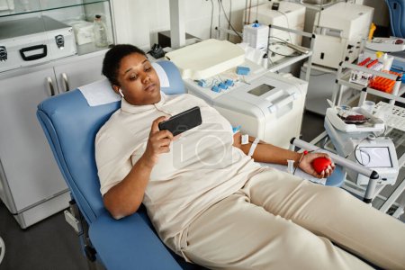 Photo for High angle portrait of black woman giving blood while laying in chair at blood donation center and holding smartphone, copy space - Royalty Free Image