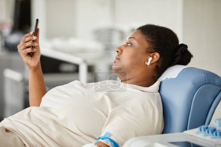 Foto de Vista lateral de la joven negra donando sangre mientras está sentada en la silla en el centro de donación de sangre y sosteniendo el teléfono inteligente - Imagen libre de derechos