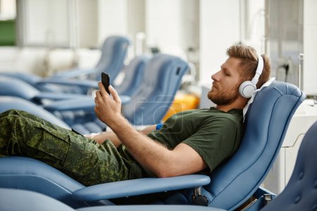 Photo for Side view portrait of man donating blood in comfort and watching videos via smartphone - Royalty Free Image