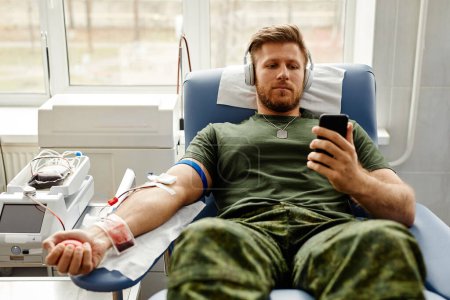 Photo for Portrait of young military man donating blood with comfort and watching videos at smartphone, copy space - Royalty Free Image