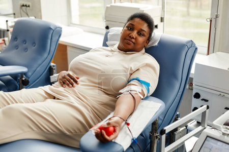 Téléchargez les photos : Portrait de jeune femme noire faisant don de sang dans une chaise confortable et regardant la caméra, l'espace de copie - en image libre de droit