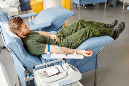 Foto de Retrato de cuerpo entero del joven soldado donando sangre mientras está acostado en una cómoda silla en el centro médico - Imagen libre de derechos