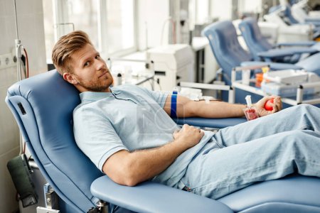 Foto de Retrato de cuerpo entero del joven donando sangre mientras está tumbado en una cómoda silla en el centro médico y mirando a la cámara - Imagen libre de derechos
