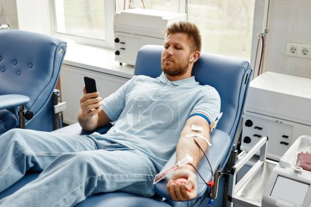 Téléchargez les photos : Portrait de jeune homme faisant don de sang tout en étant allongé dans une chaise confortable au centre médical et en utilisant un smartphone - en image libre de droit