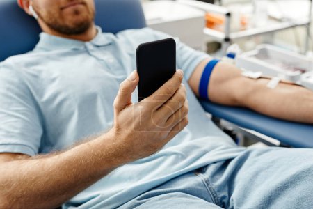 Téléchargez les photos : Gros plan d'un jeune homme qui donne du sang alors qu'il est allongé sur une chaise confortable au centre médical et tient un smartphone - en image libre de droit
