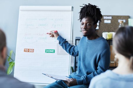 Foto de Retrato de un joven negro enseñando inglés en la oficina a un grupo de personas y señalando la pizarra blanca con reglas gramaticales - Imagen libre de derechos