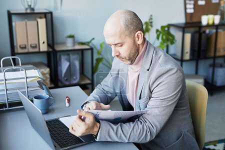 Téléchargez les photos : Portrait d'un adulte chauve lisant un document de données et utilisant un ordinateur portable tout en travaillant dans un bureau d'entreprise - en image libre de droit