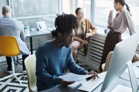 Téléchargez les photos : Portrait de jeune homme noir utilisant un ordinateur portable sur le lieu de travail avec des gens bavardant en arrière-plan dans le bureau de l'entreprise, espace de copie - en image libre de droit