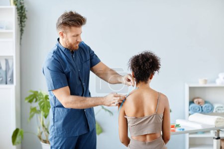 Photo for Side view portrait of rehabilitation therapist putting physio tape on shoulder of young woman to relieve muscle pain, copy space - Royalty Free Image
