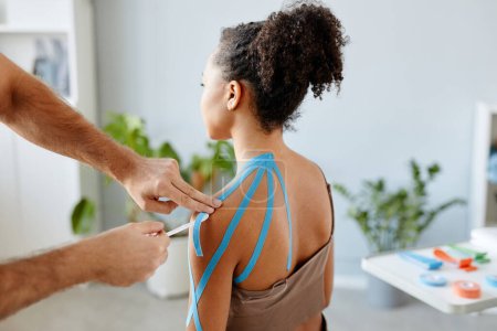 Photo for Close up rehabilitation therapist putting k tape on shoulder of young African American woman in physiotherapy clinic - Royalty Free Image