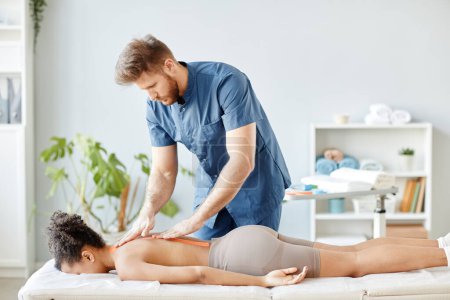 Photo for Portrait of rehabilitation specialist putting physio tape on back of young woman lying on massage bed - Royalty Free Image
