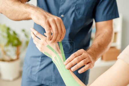 Closeup rehabilitation therapist putting physio tape on hand and arm of young woman to relief joint pain, copy space
