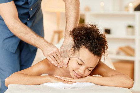 Téléchargez les photos : Vue de face portrait de jeune femme noire bénéficiant d'un massage du dos dans une séance de SPA relaxant et souriant avec bonheur - en image libre de droit