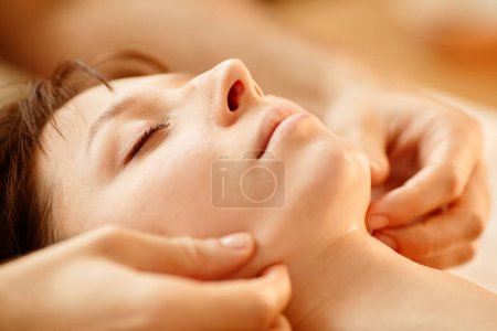 Foto de Cálido primer plano tonificado de la mujer joven disfrutando de la sesión de masaje facial en el centro de SPA con los ojos cerrados - Imagen libre de derechos