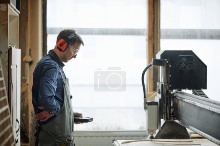 Téléchargez les photos : Portrait latéral du menuisier masculin utilisant une machine de découpe CNC dans un atelier de production automatisé - en image libre de droit