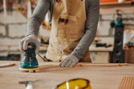 Foto de Primer plano de carpintero joven que lija madera mientras que construye muebles hechos a mano en taller, espacio de la copia - Imagen libre de derechos