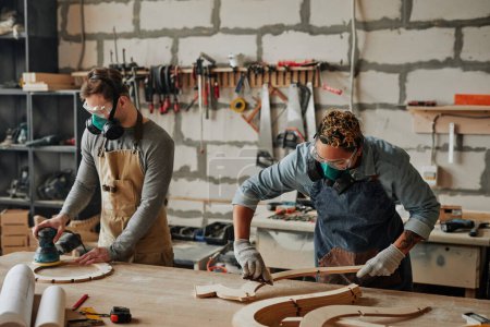 Téléchargez les photos : Portrait chaleureux de deux menuisiers en équipement de protection travaillant avec du bois tout en construisant des meubles artisanaux en atelier - en image libre de droit
