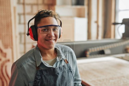 Photo for Portrait of female worker smiling at camera while wearing noise cancelling headphones in workshop, copy space - Royalty Free Image