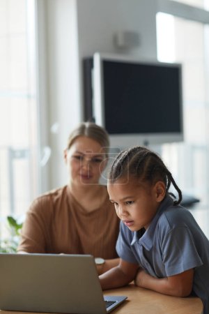 Téléchargez les photos : Portrait vertical de mignonne fille noire regardant l'écran d'ordinateur portable avec une mère caucasienne souriante en arrière-plan - en image libre de droit