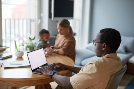 Photo for Side view portrait of black man using laptop at home with wife and children in background, copy space - Royalty Free Image