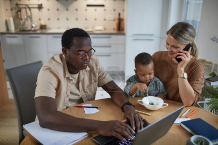Téléchargez les photos : Portrait de père noir travaillant à la maison avec sa femme et son enfant assis à table à côté de lui - en image libre de droit