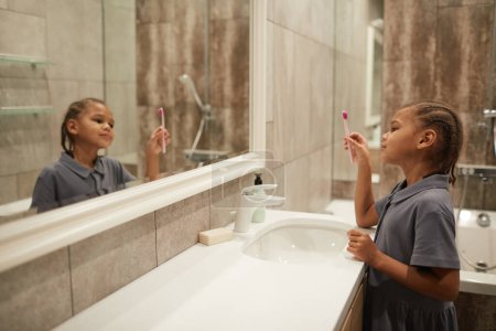 Photo for Side view portrait of cute black girl holding toothbrush and looking at mirror in bathroom learning to practice dental hygiene, copy space - Royalty Free Image