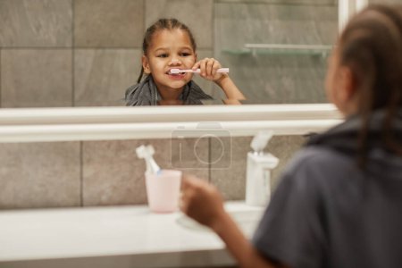 Photo for Close up portrait of cute black girl brushing teeth and looking in bathroom mirror, copy space - Royalty Free Image