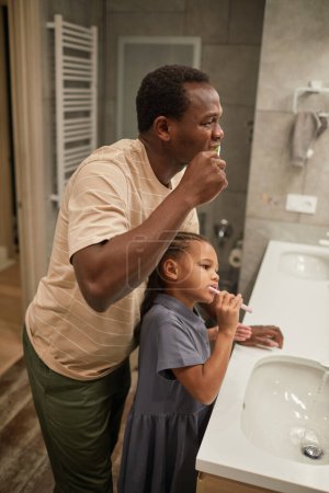 Téléchargez les photos : Vue de côté portrait de père noir et fille brossant les dents ensemble et regardant miroir dans la salle de bain - en image libre de droit