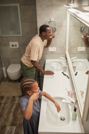 Foto de Alto ángulo de padre negro e hija cepillándose los dientes juntos y mirando al espejo en el baño - Imagen libre de derechos
