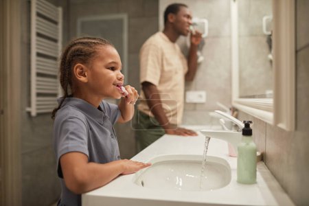 Foto de Vista lateral de linda chica negra cepillándose los dientes con papá y mirando al espejo en el baño - Imagen libre de derechos