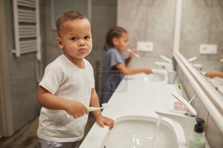 Photo for Portrait of two African American children in bathroom focus on little toddler boy brushing teeth and looking at camera, copy space - Royalty Free Image